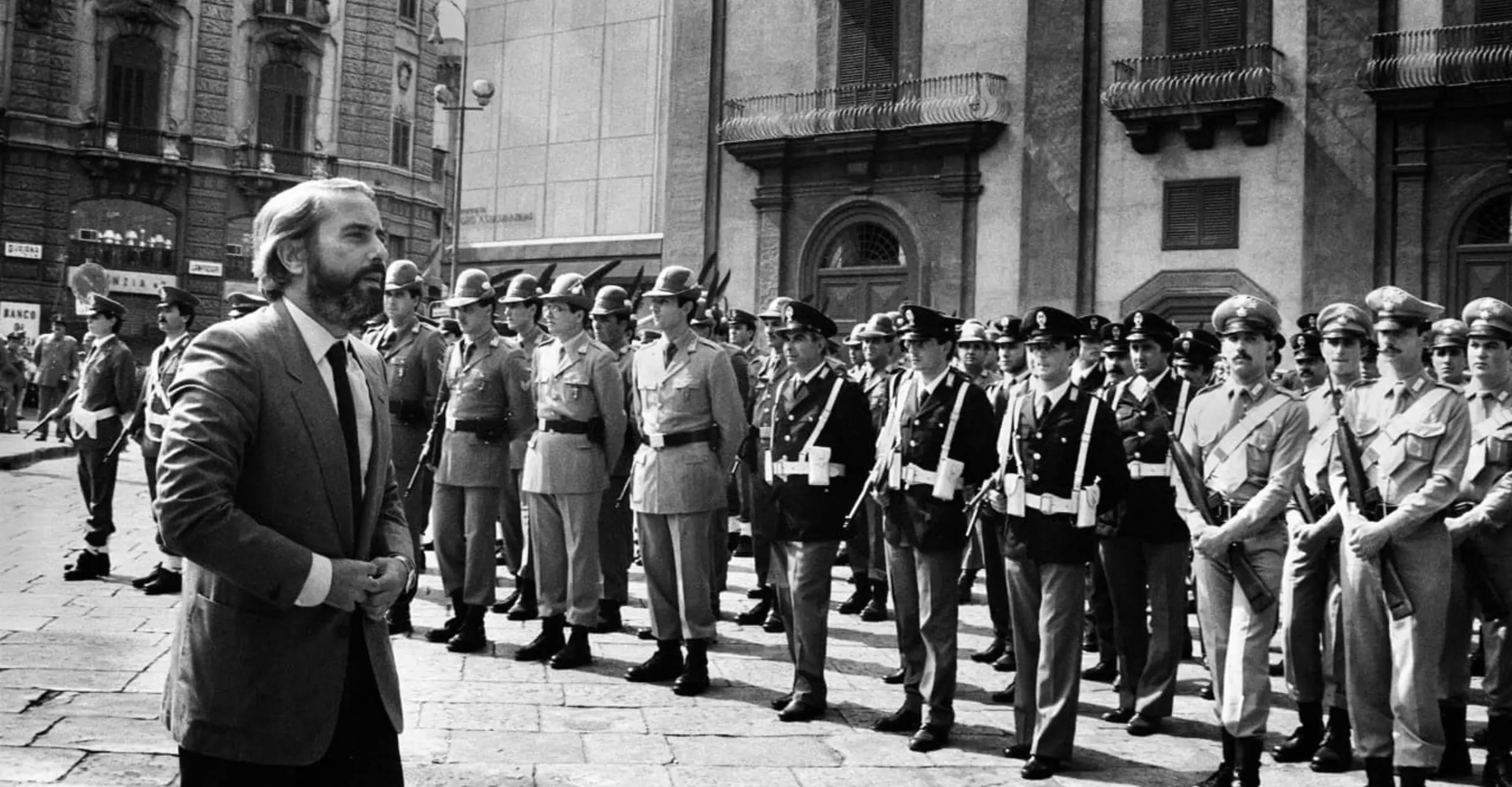 Black and white photograph of a suited man walking past a military parade. 