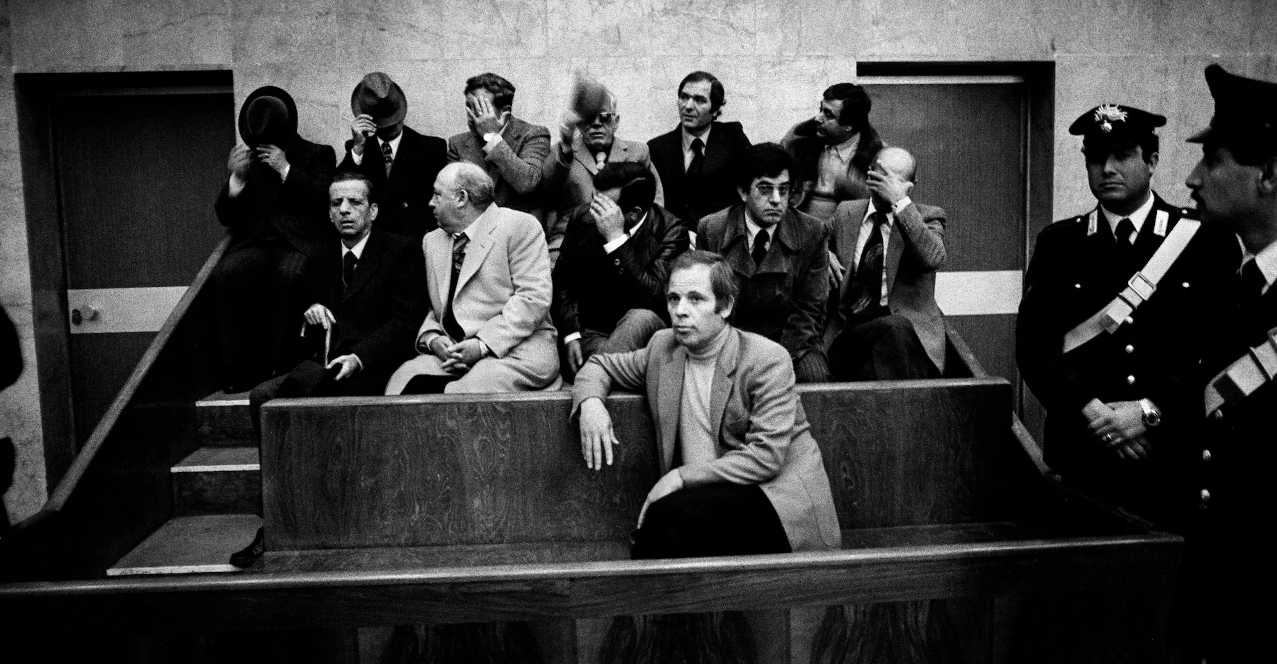 Black and white photograph of a man sat in a relaxed position in a court dock, flanked by authority figures. Several men are sat behind him attempting to hide their faces.