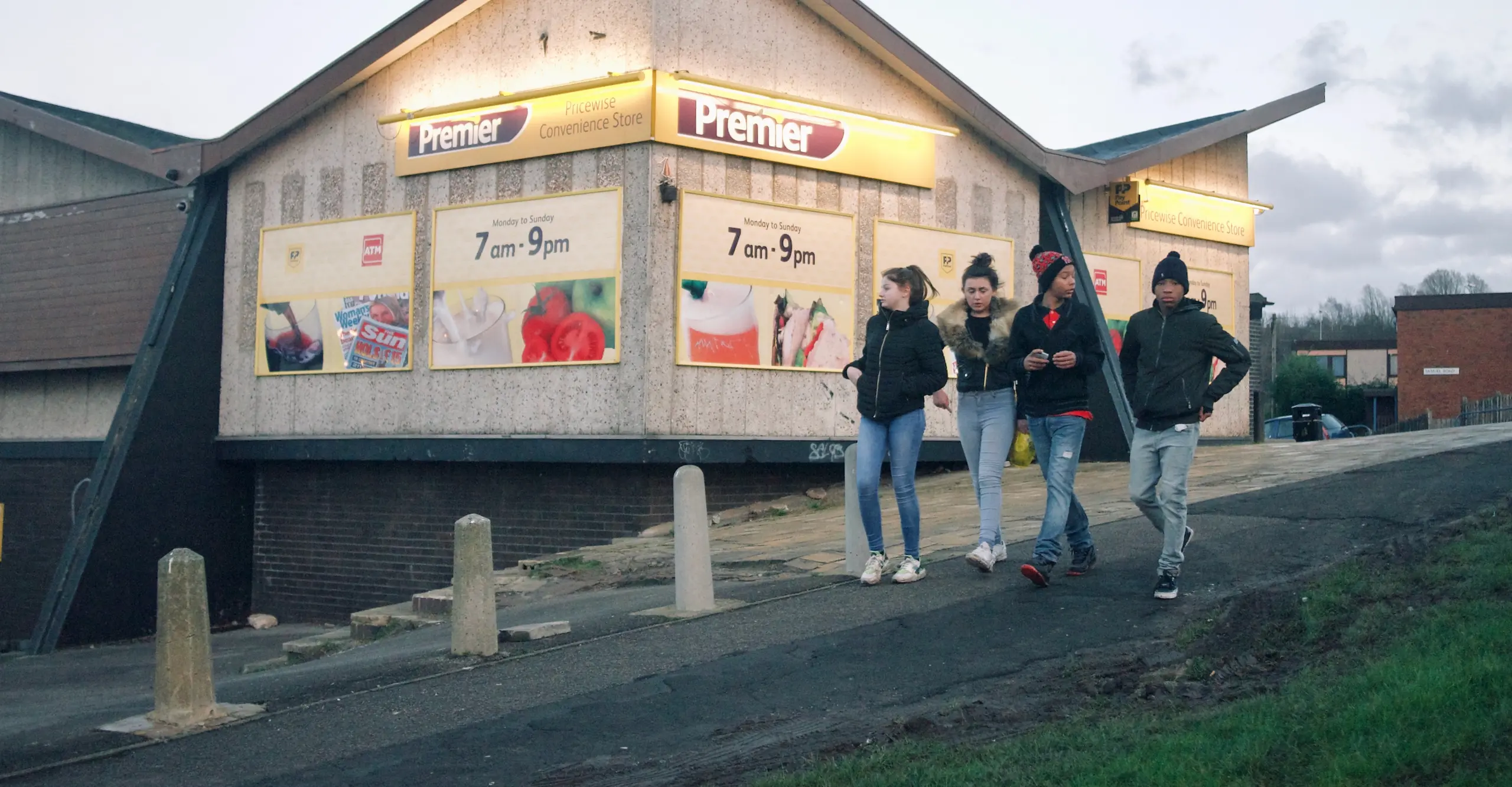 Four young people walk in a row down a paved hill past a Premier convenience store at dusk.