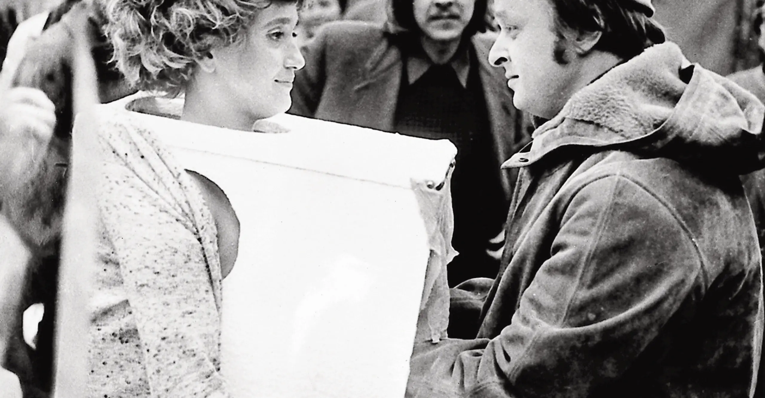 Black and white photo of a woman having her breasts in a box with a curtain felt by a man.