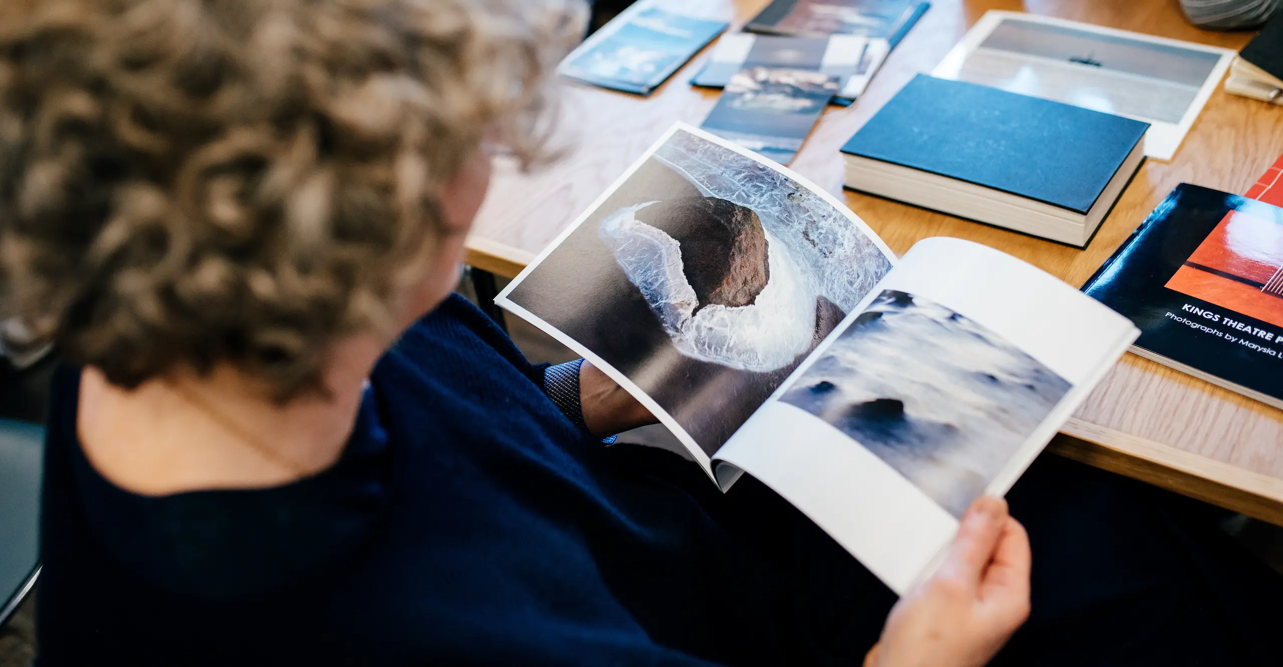 Over the shoulder photo of someone seated at a table looking at a photography book showing seascape photographs.