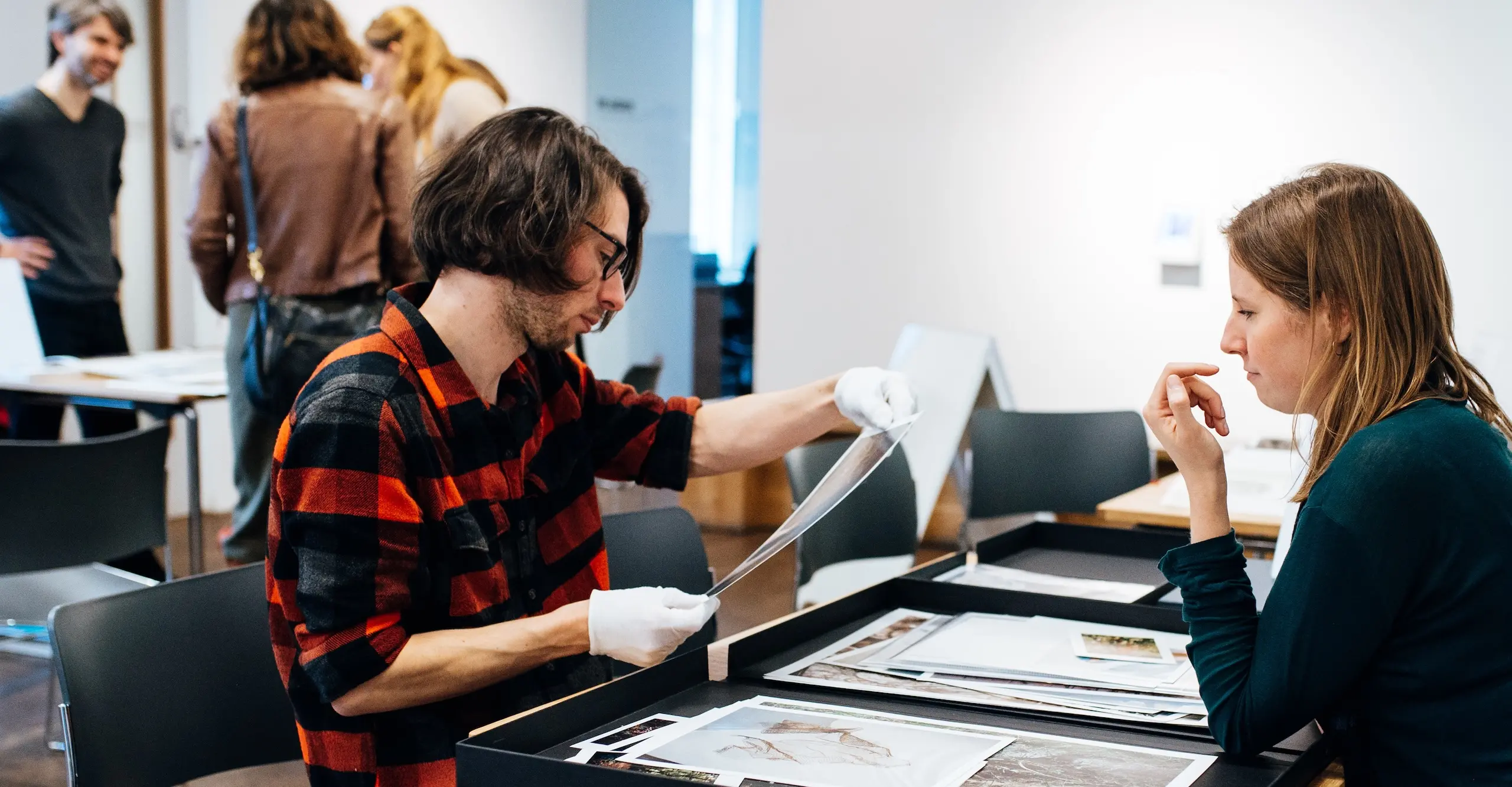 Colour photograph of two people sitting across from each other at a table with photographs laid out on it