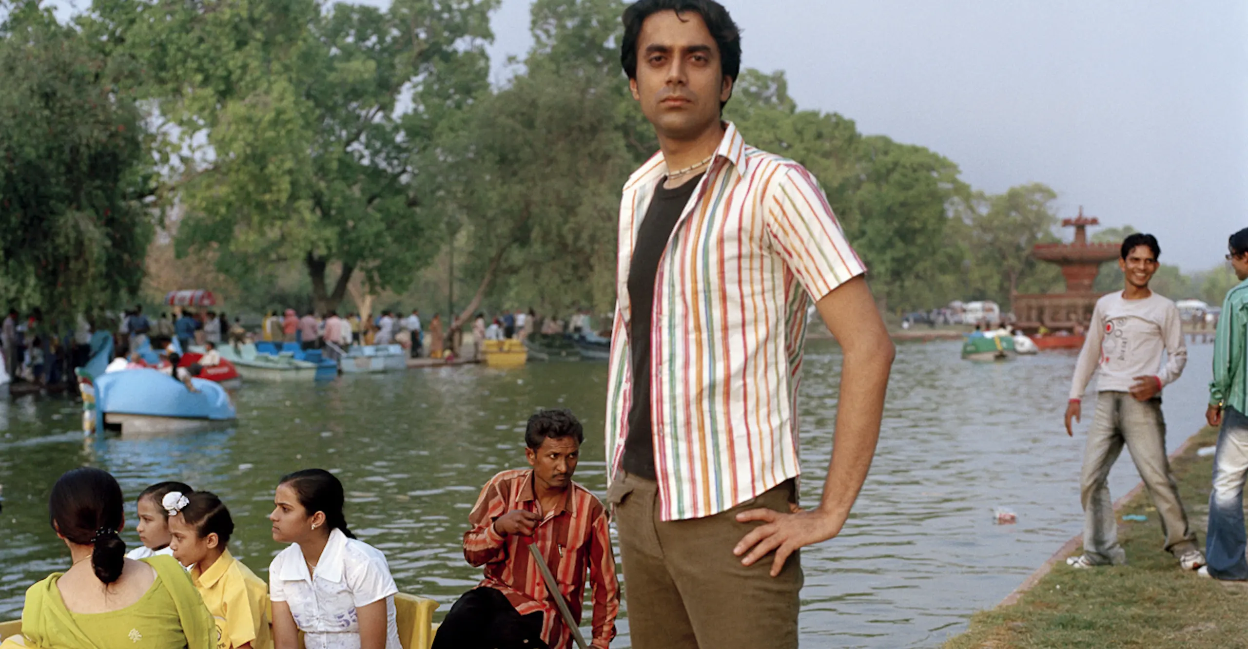 Figure standing on a riverbank looking to camera as a small pleasure boat floats alongside.