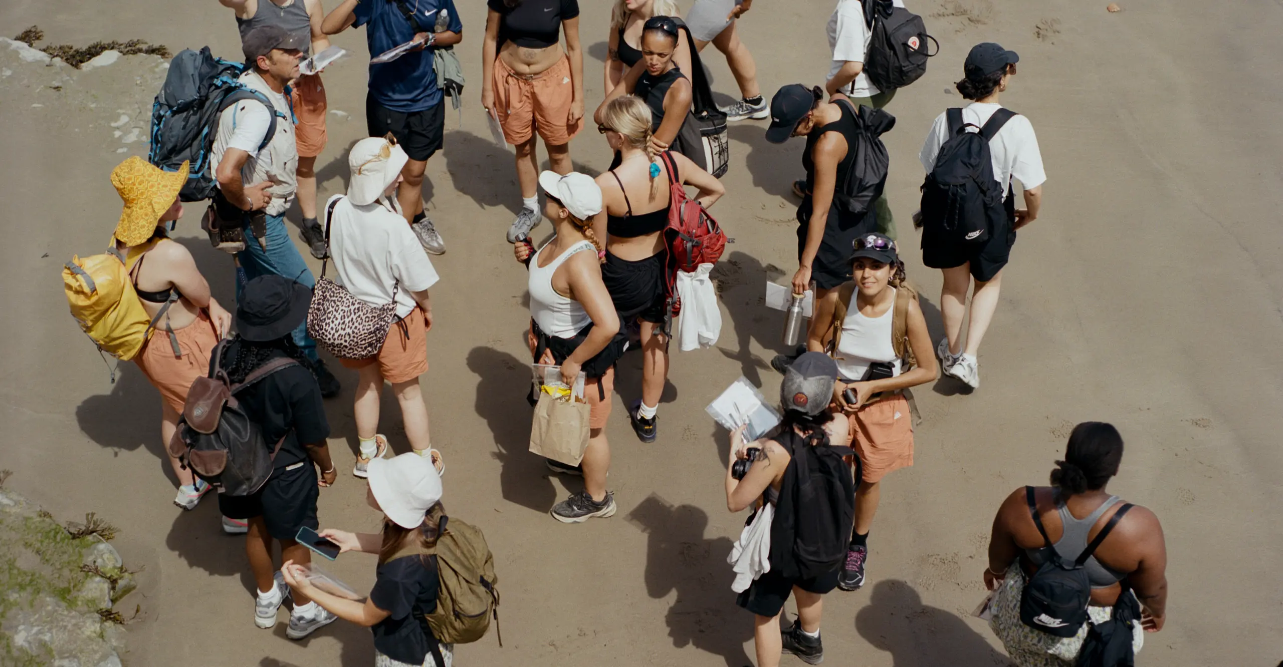 Arial view of a group of mostly women standing in a group on a beach wearing hiking gear.