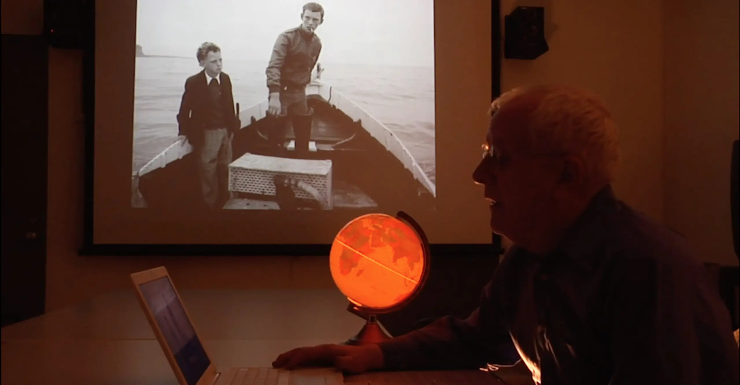 Chris Killip sits at a desk in profile, a black and white photo is projected beyond him. 