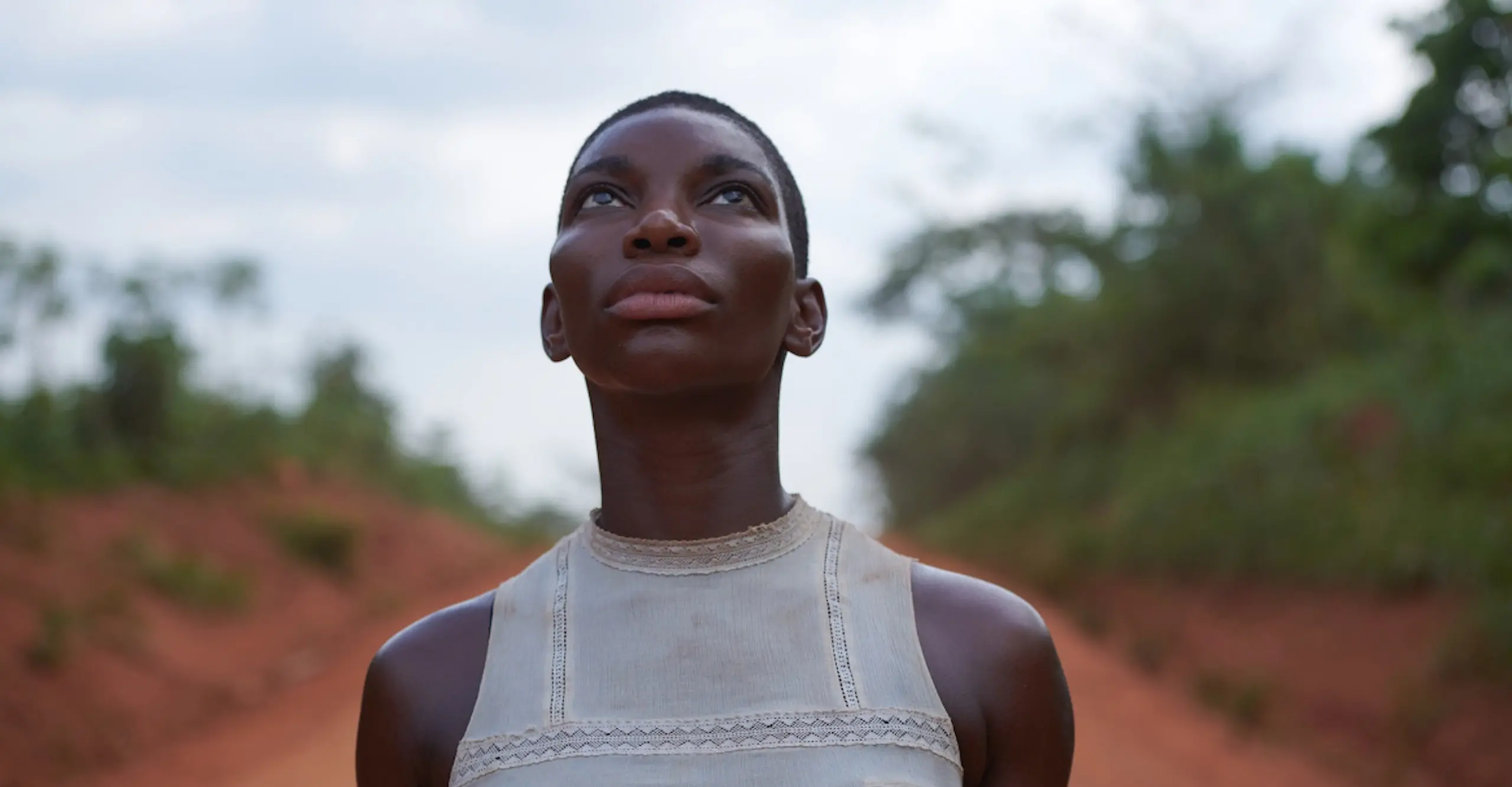 A woman stands on a red dirt road, face turned upward, wearing a sleeveless, cream muslin shirt.