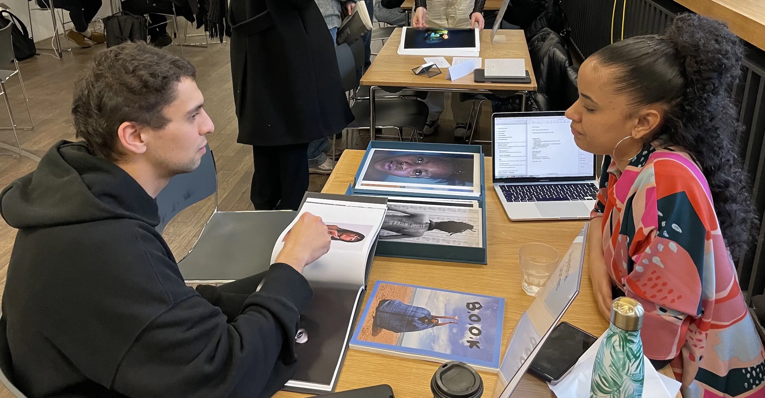 Two people sit cross from each other at a table with photo publications in front of them. 