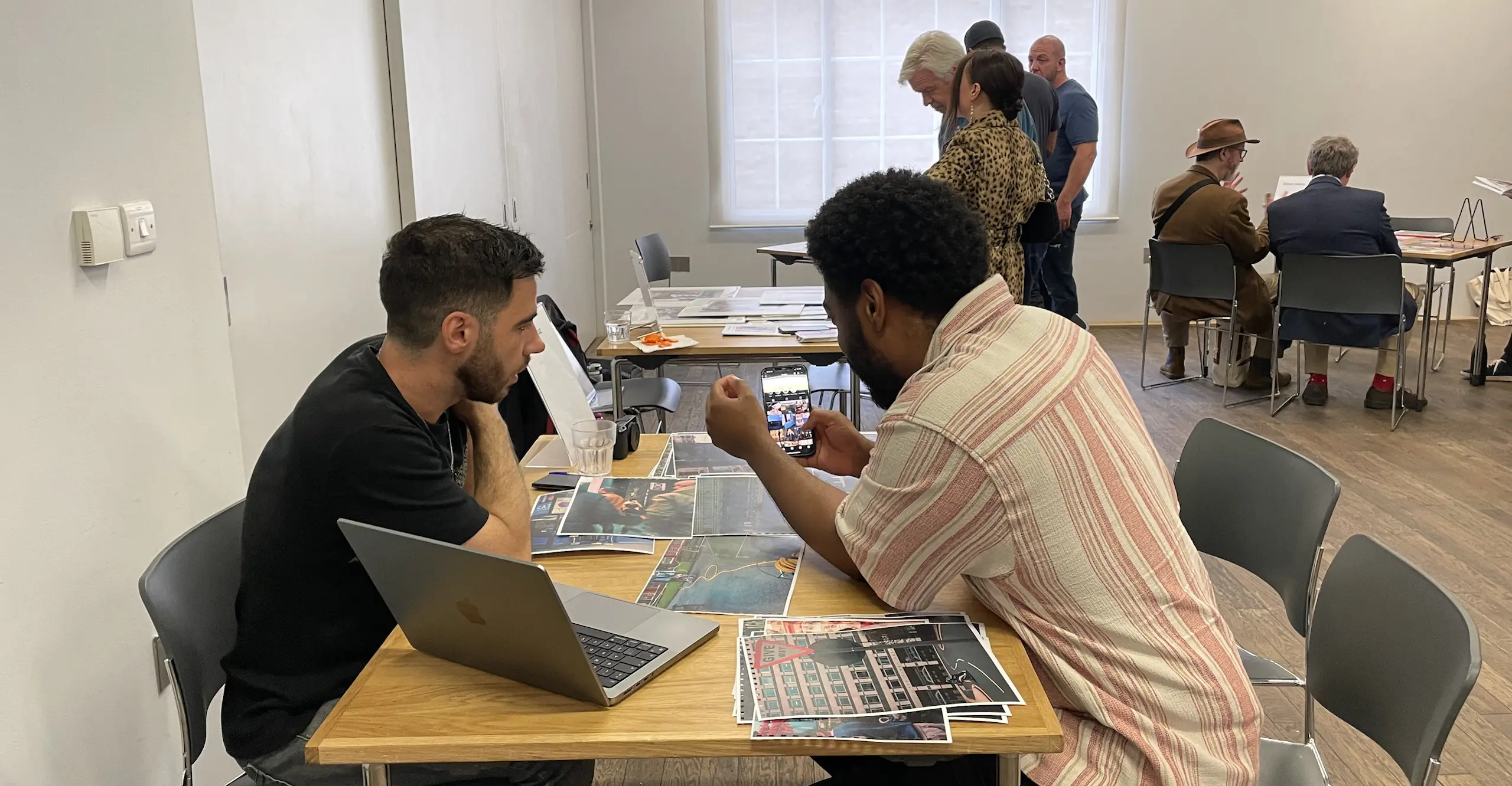 Two people sit opposite each other at a table with photographs spread across it, other people are at tables in the background.