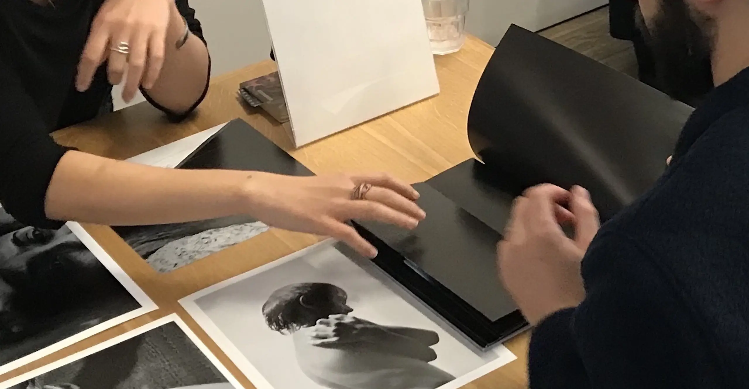 A table top with black and white photos on it, with the hands and arms of two people sitting across from each other.