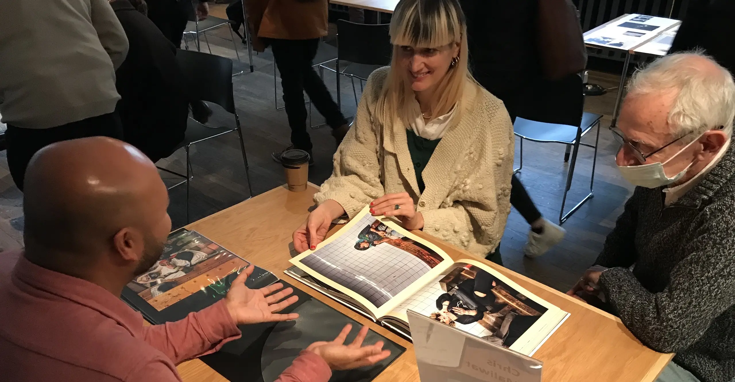 Two people sit at a table opposite another person who his presenting his photographs on the table