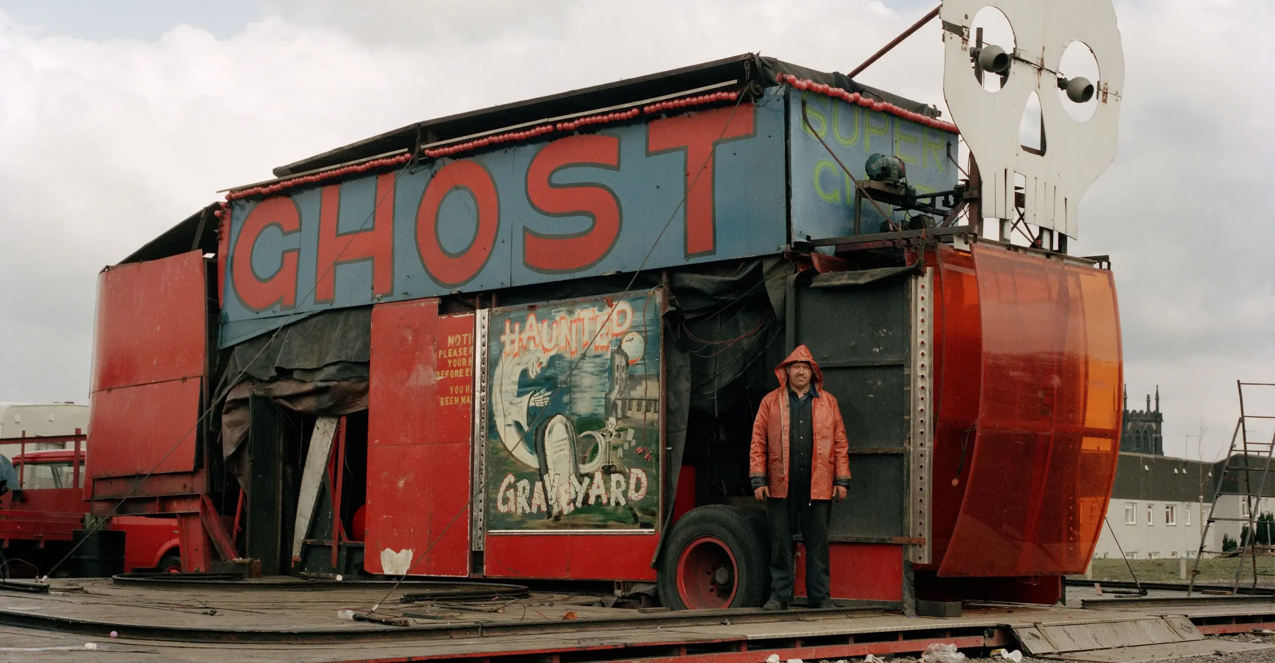 Colour photograph of a man standing in front of a packed up fairground ride called Ghost Train.