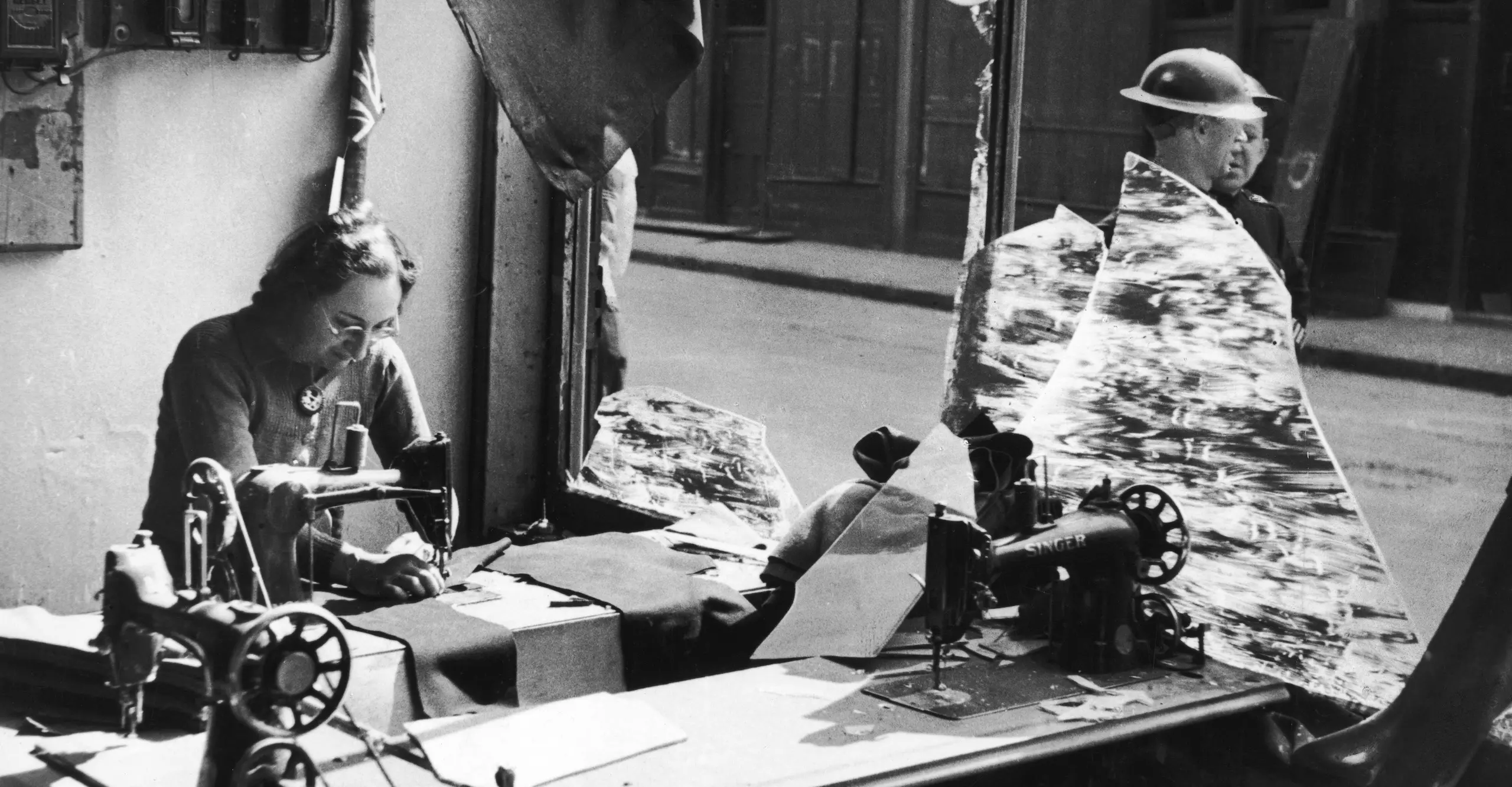 Black and white photograph of a woman working amongst the broken glass of a tailor’s shop.