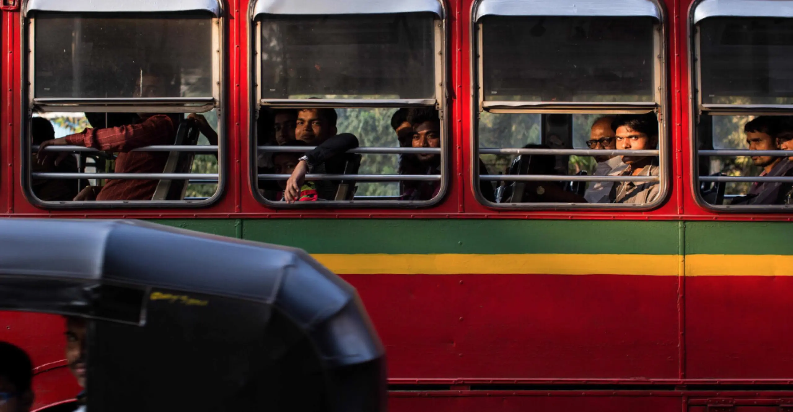 Coloured photograph featuring a bus with commuters visible through open  windows. The bus is partially obscured on the bottom right by the top of a black bike carriage