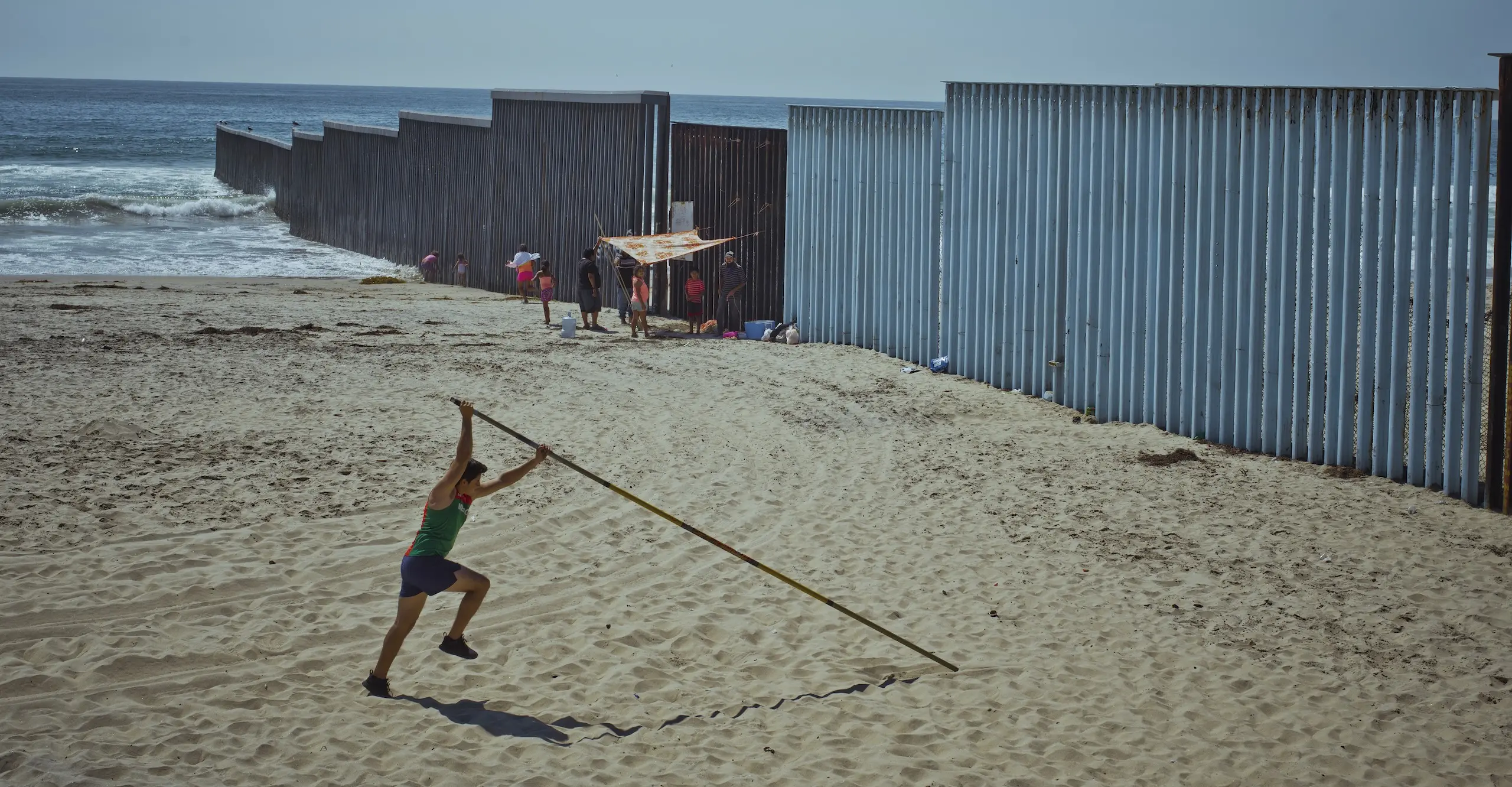 Colour photograph of a beach with a person holding a long stick outstretched above their head simulating a high jum[er