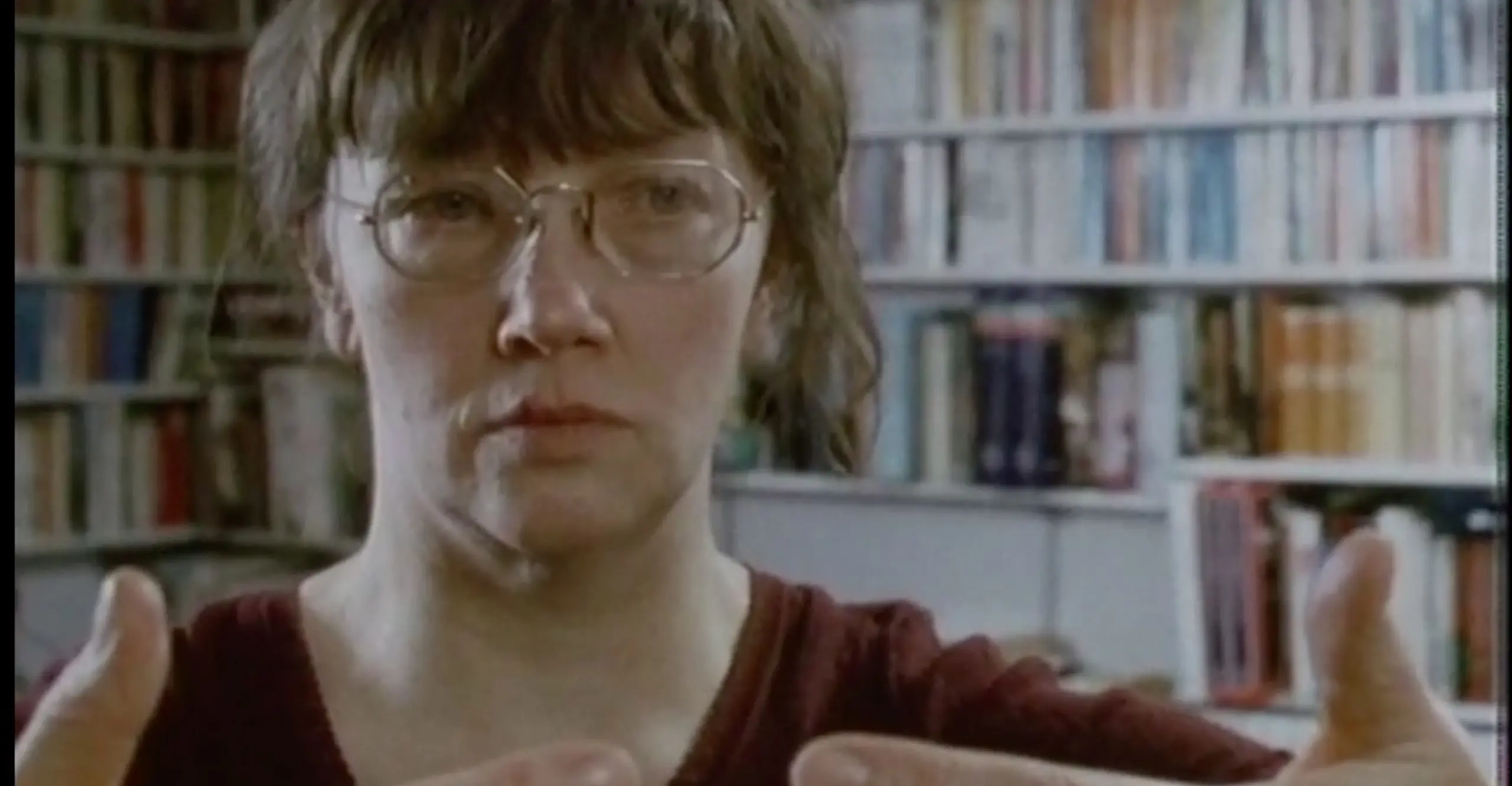 A white woman with glasses and brown hair holds her hands up in front of her, fingers framing under her neck and face.