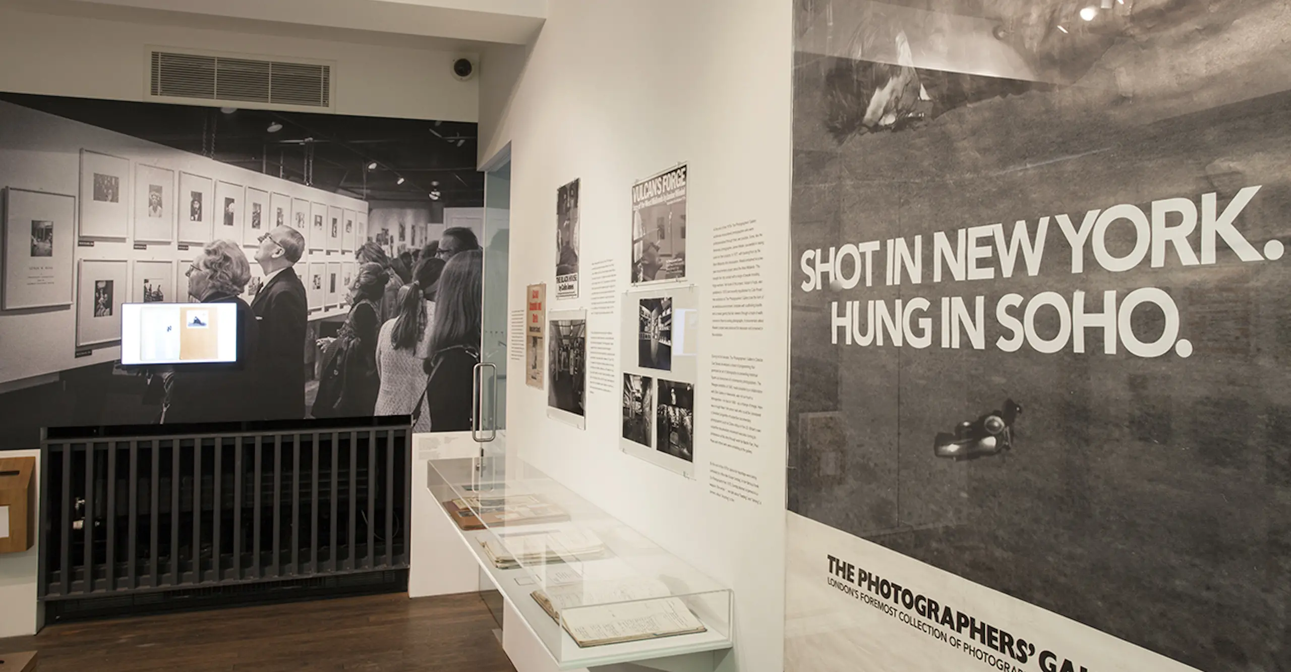 Exhibition room displaying a poster, photographs and text on a wall and a vitrine with open books inside.