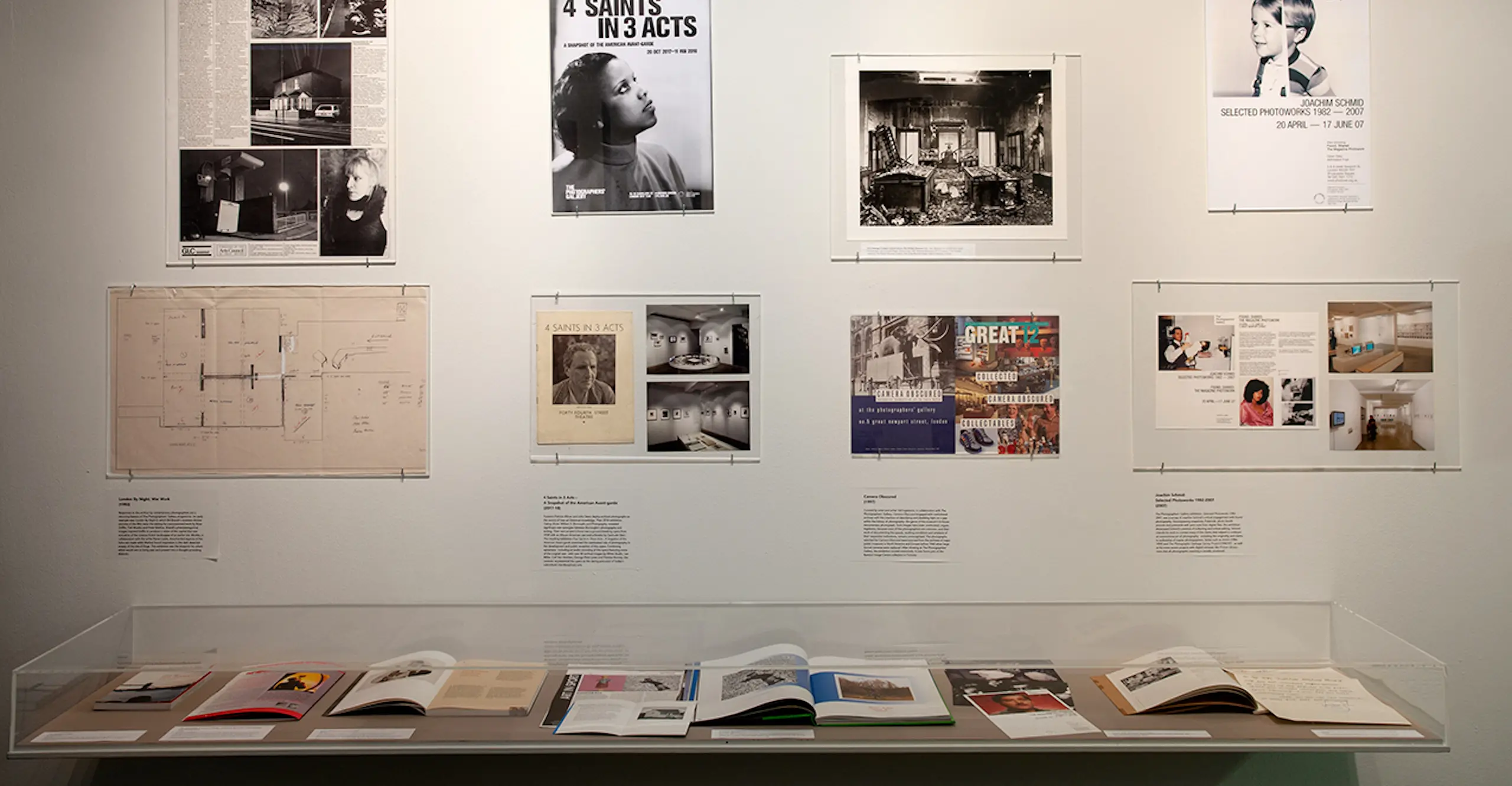 A gallery wall with photographs, posters and writing underneath which sits a vitrine with open books and magazines on display.