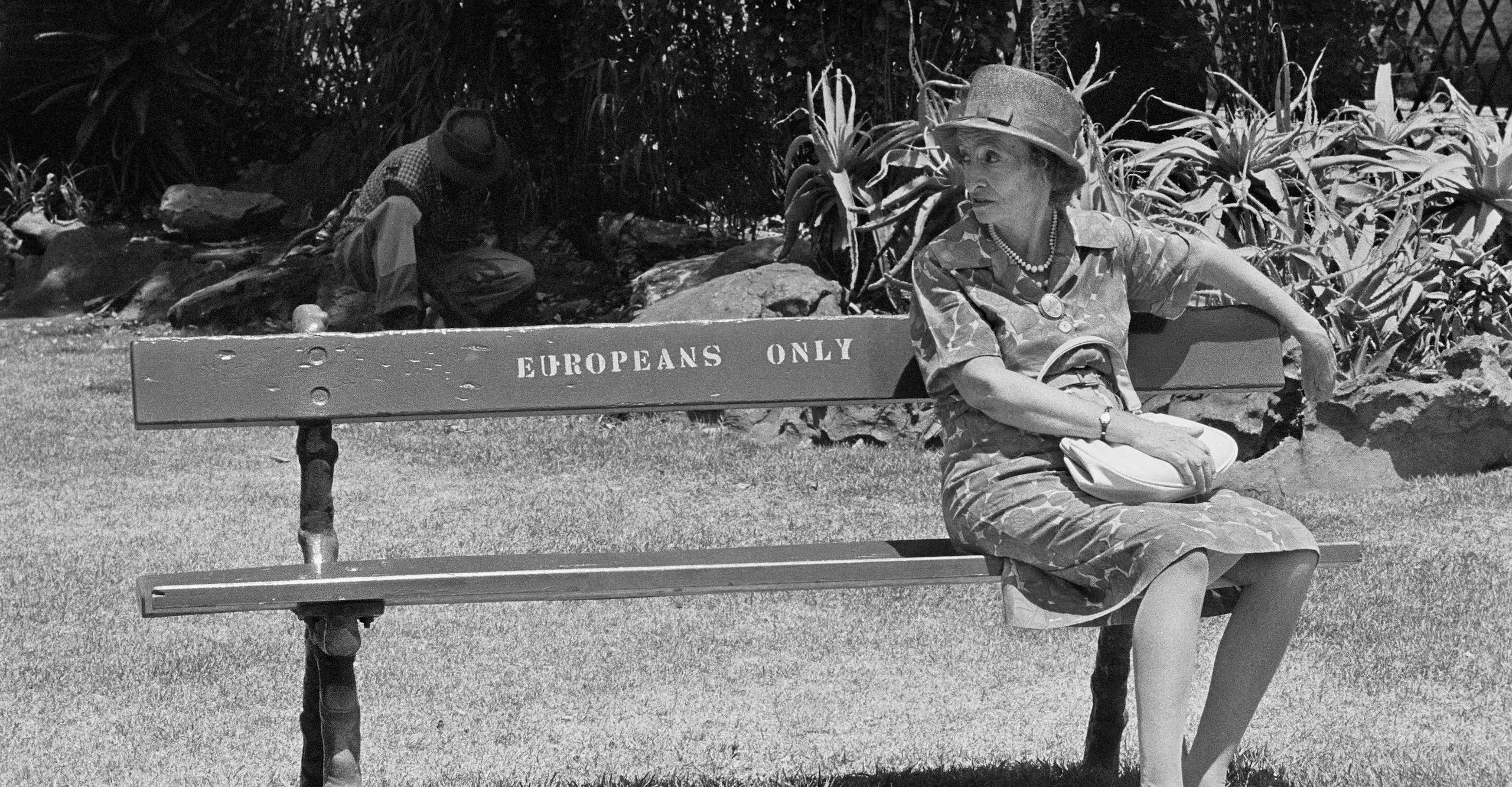 Black and white photograph of a white woman sat alone on an outdoor bench with the markings “EUROPEANS ONLY.”