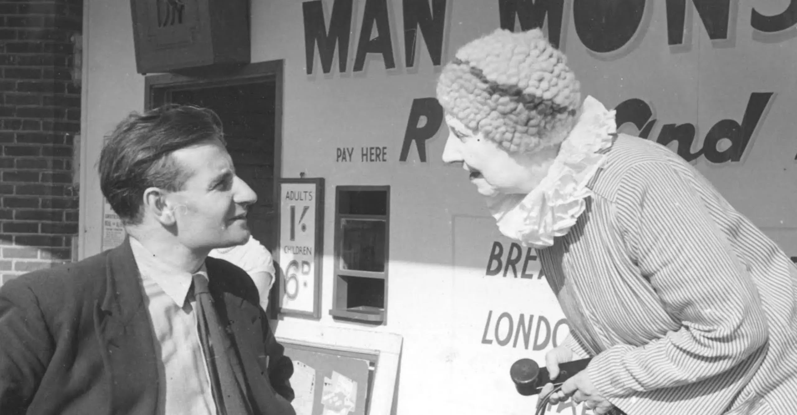 Two people stand in a fairground, one higher up and leaning down to speak with the other.