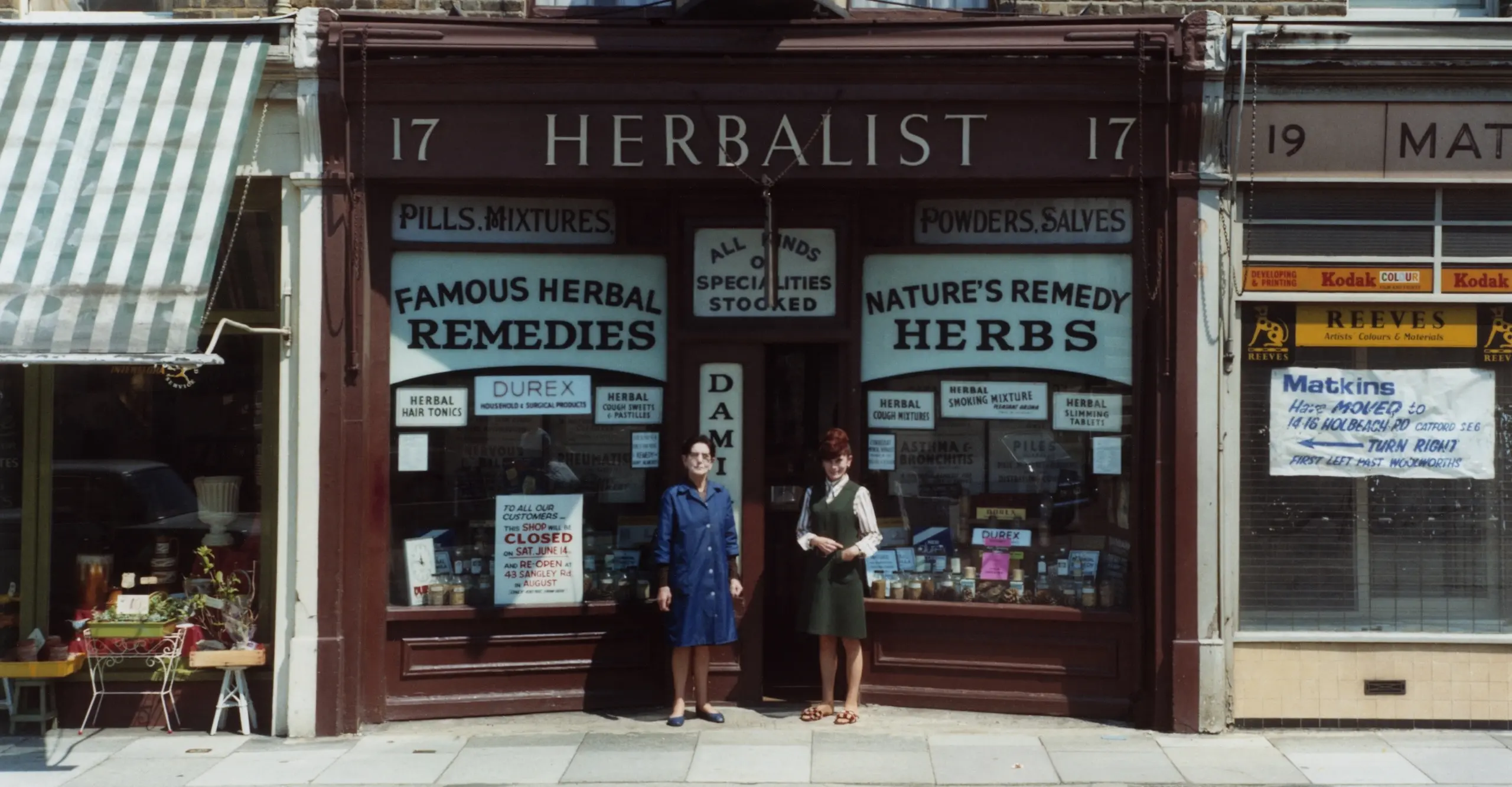 A colour photograph of a 'Herbalist' storefront. Two smartly dressed women are pictures standing in front of the shop.