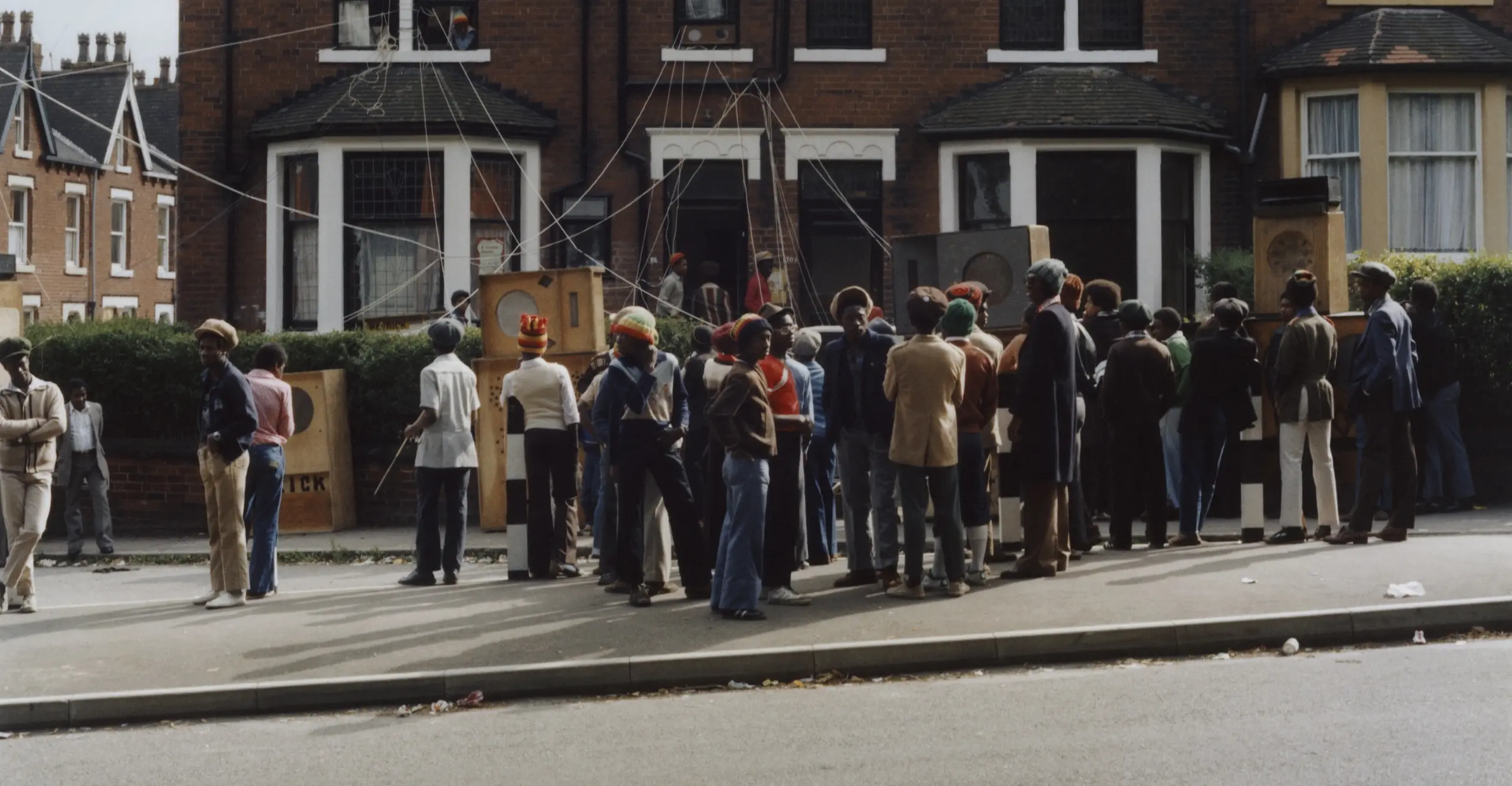 Colour photograph taken across the street from a house with a group of people setting up a sound system 