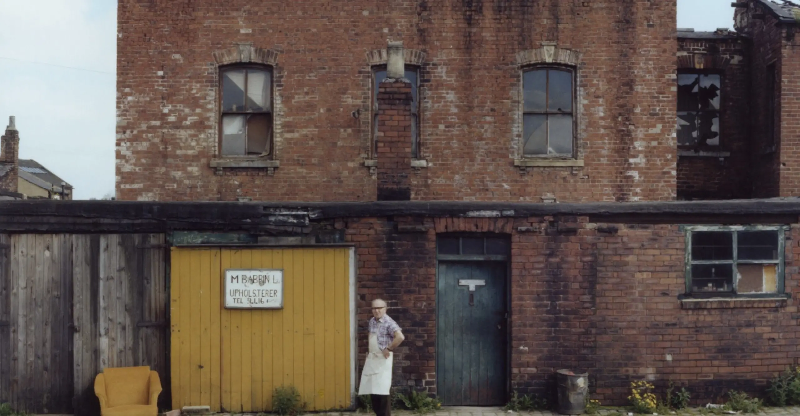 Colour photograph of the back of a house on a street. A man is standing in an apron on the street.