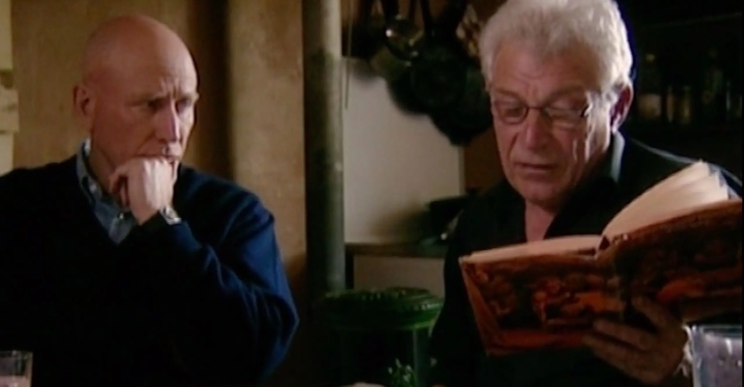 Two white men sit next to each other at at table, one reading from an open book.