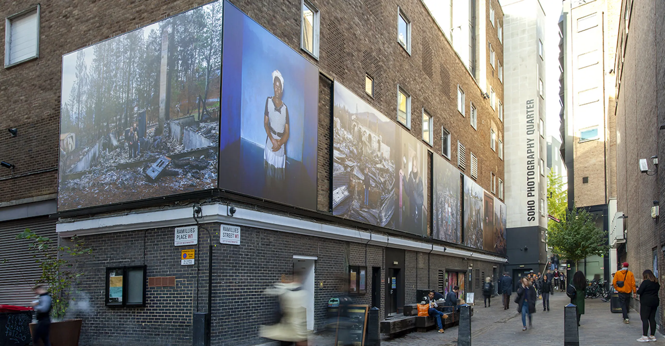 A frieze of colour photographs on an outdoor building depicting portraits of people standing in the aftermath of a fire or flood.