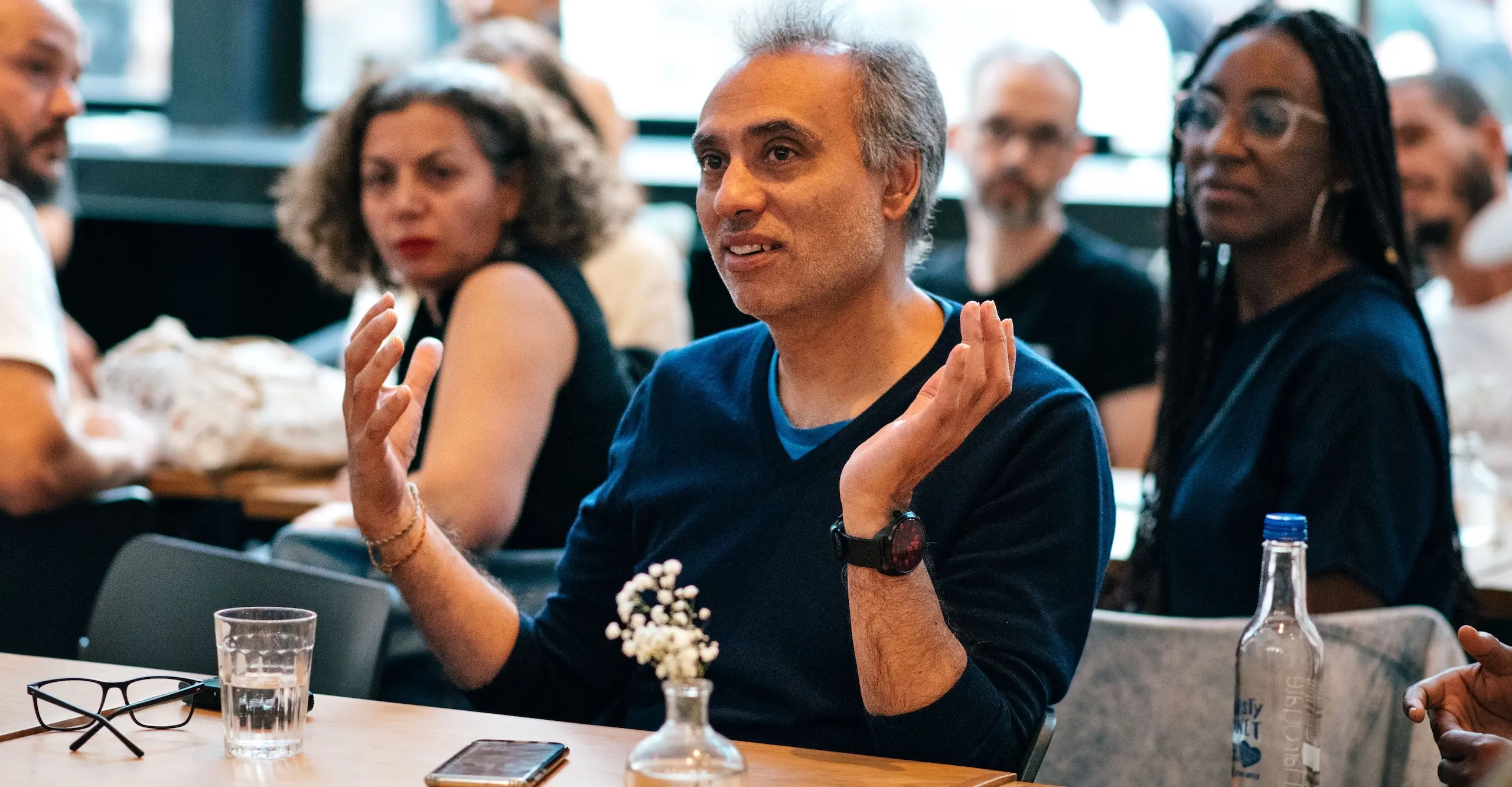 Colour photograph of people sitting at tables listening to one seated person speaking amongst them