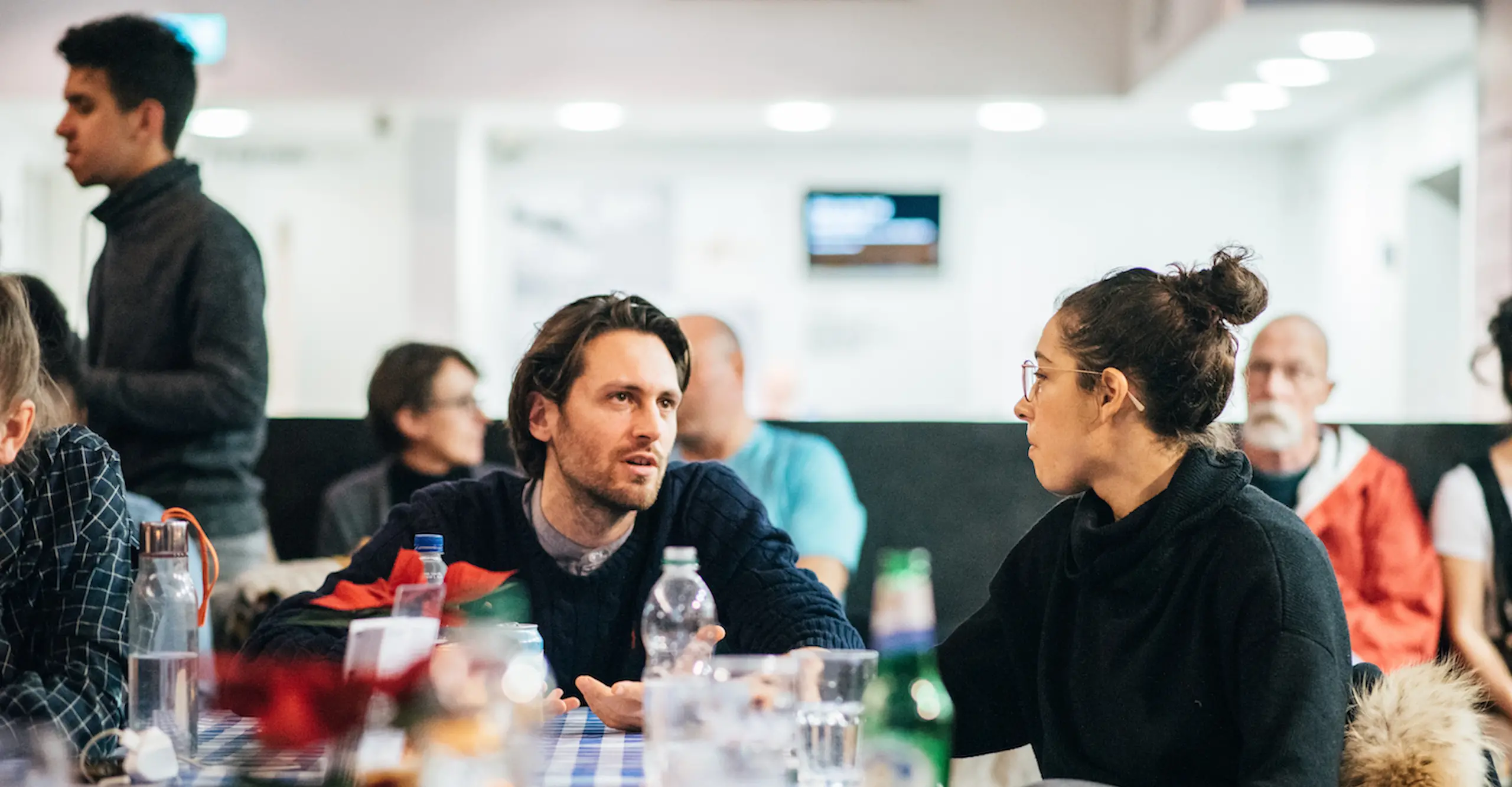 People sit and stand together in different combinations at a social gathering inside a bright space.