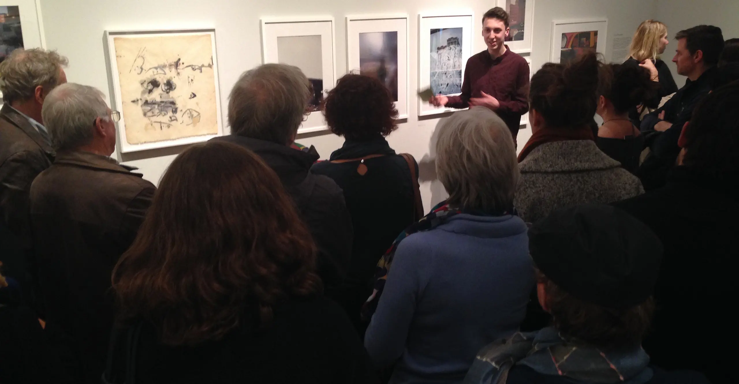 A photograph of the backs of a crowd looking at photographs on a white wall. 