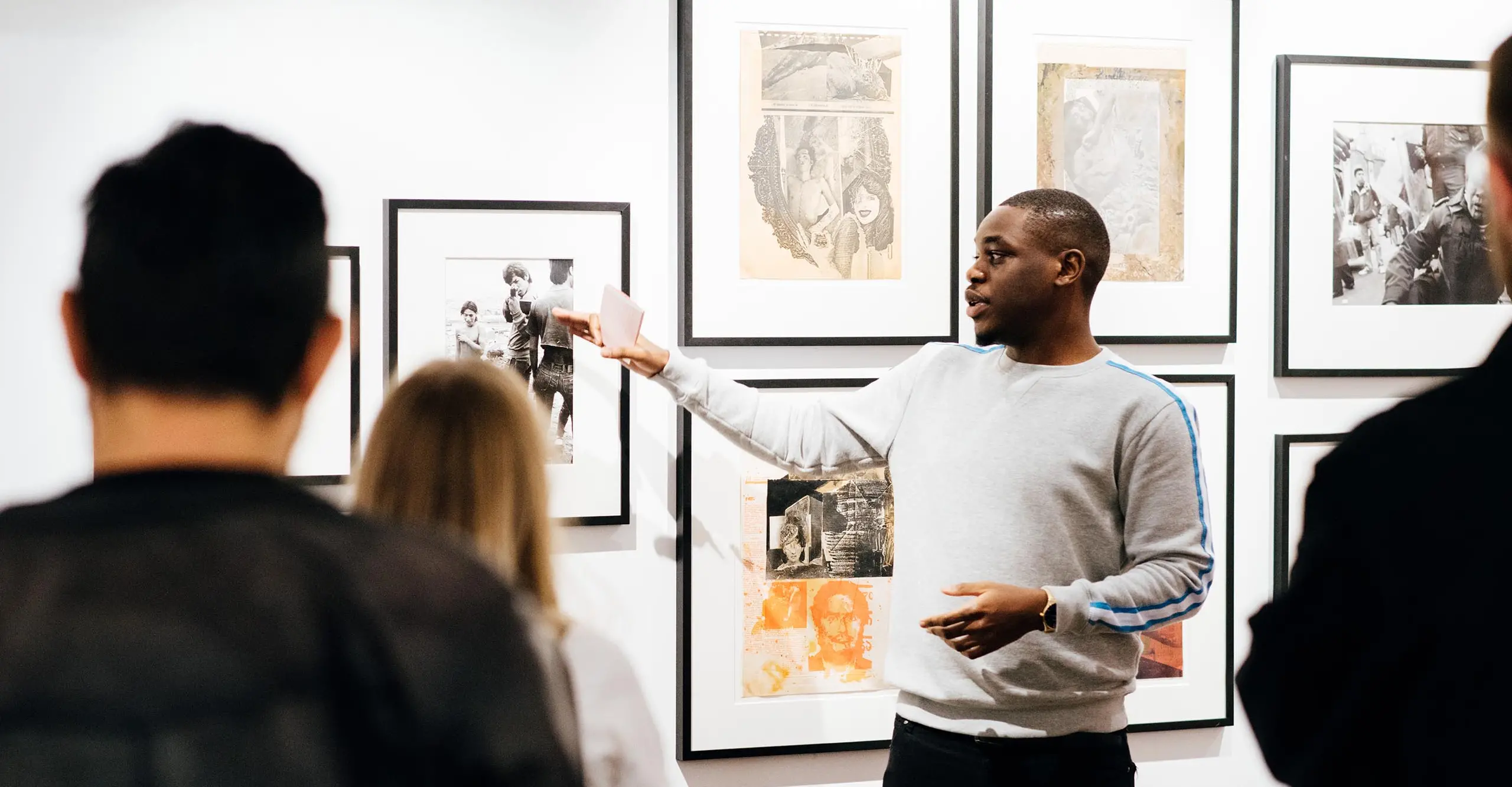 A photograph of a person in the middle of the frame pointing to a photograph on a gallery wall.