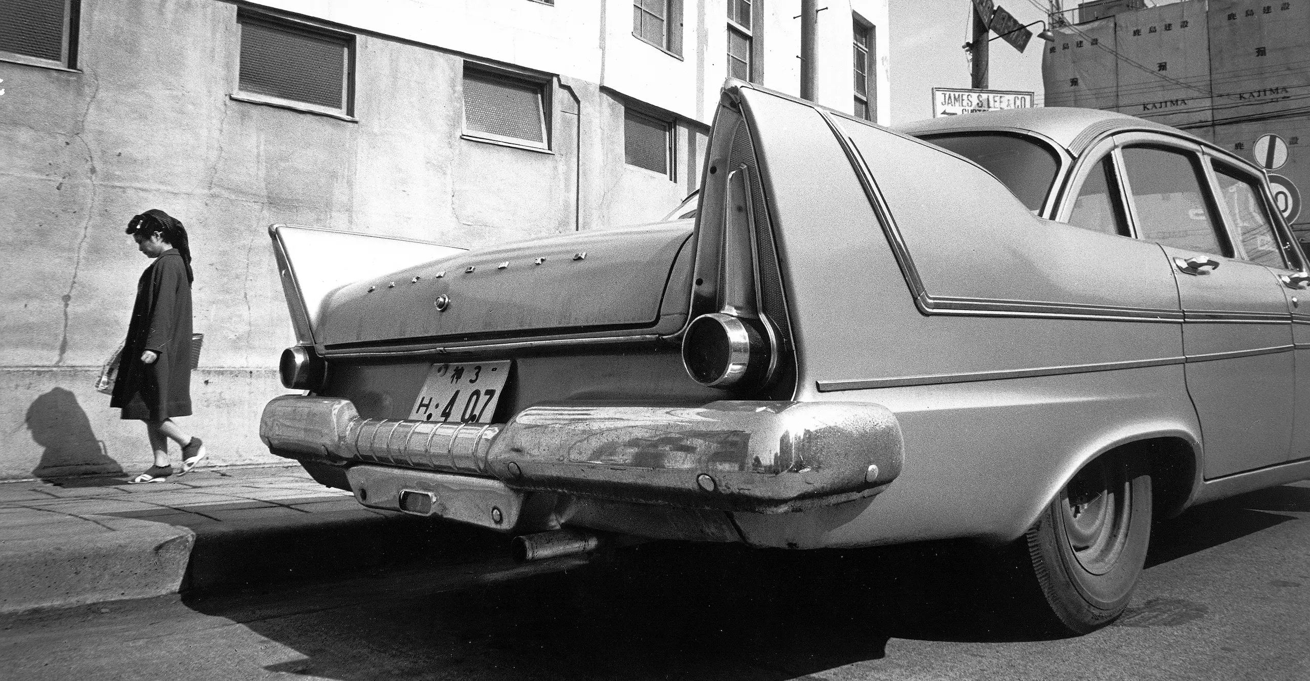 Black and white photograph of the rear of a 1960s car on a street with a person walking past