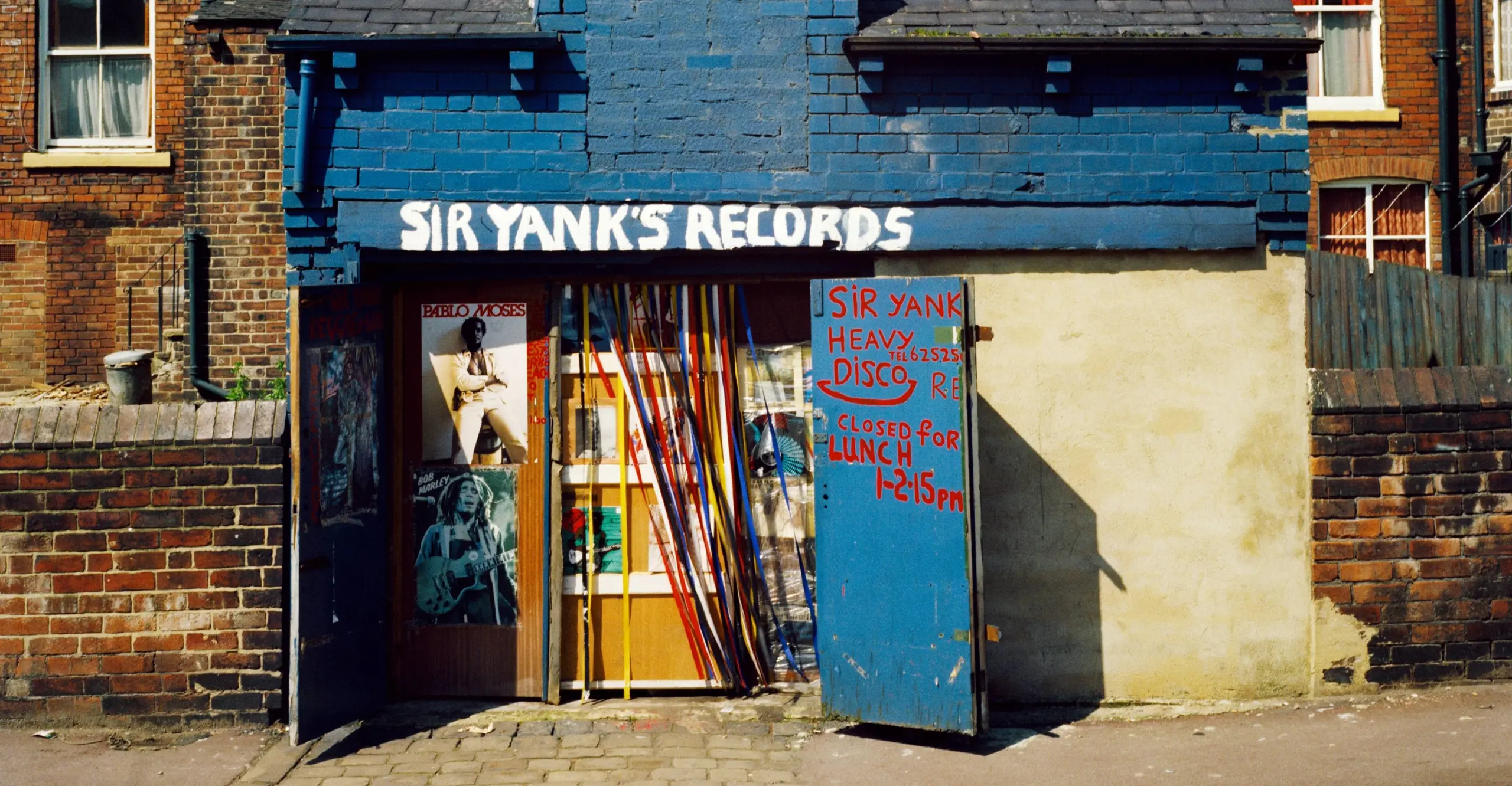 Colour photograph of a blue brick building with the  words ‘Sir Yank’s Records’ painted on. Posters of Bob Marley and Pablo Moses can be seen stuck on the interior