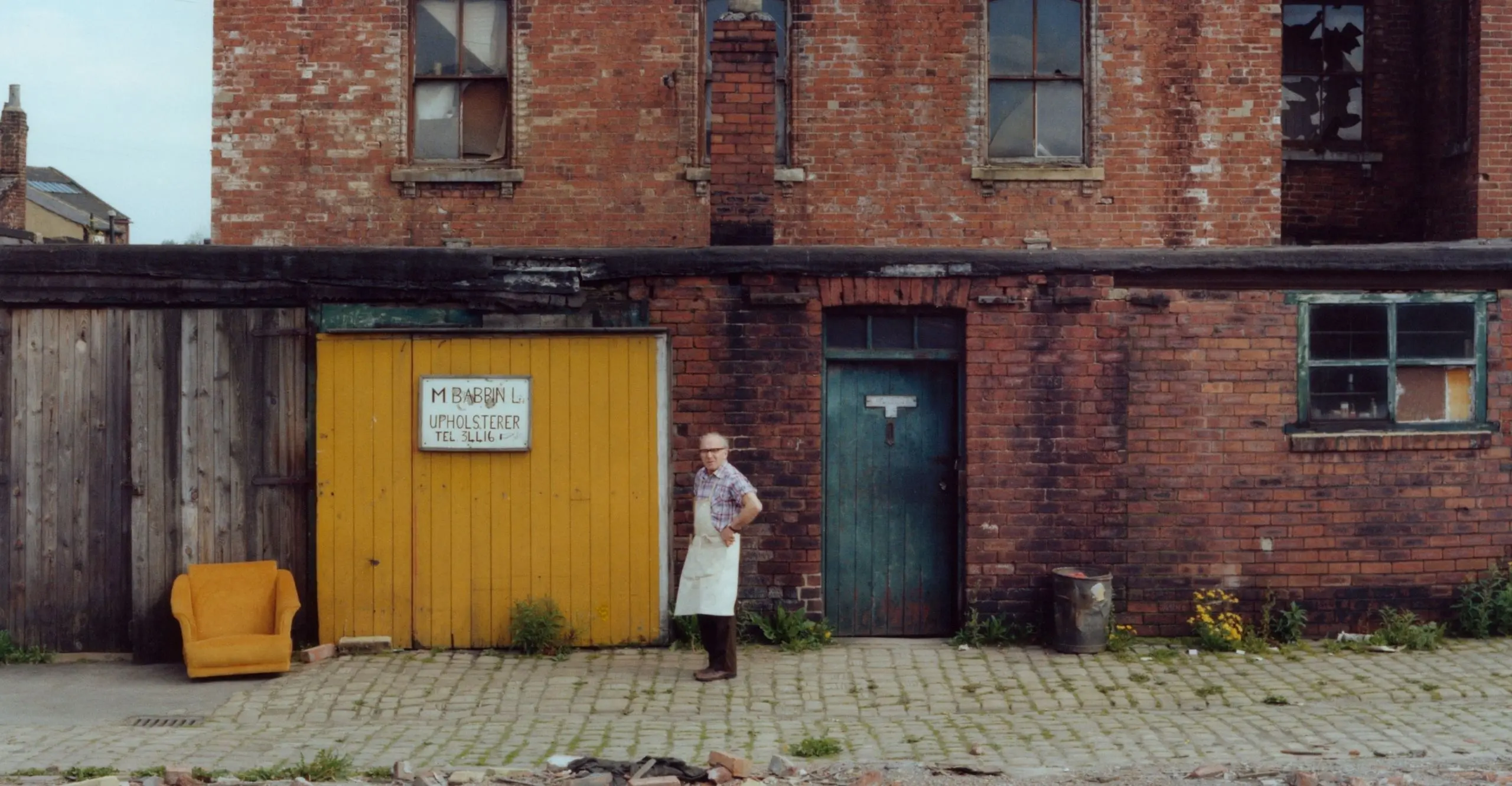  Colour photograph of the back of a house on a  street. An old man in an apron stands on the street. 