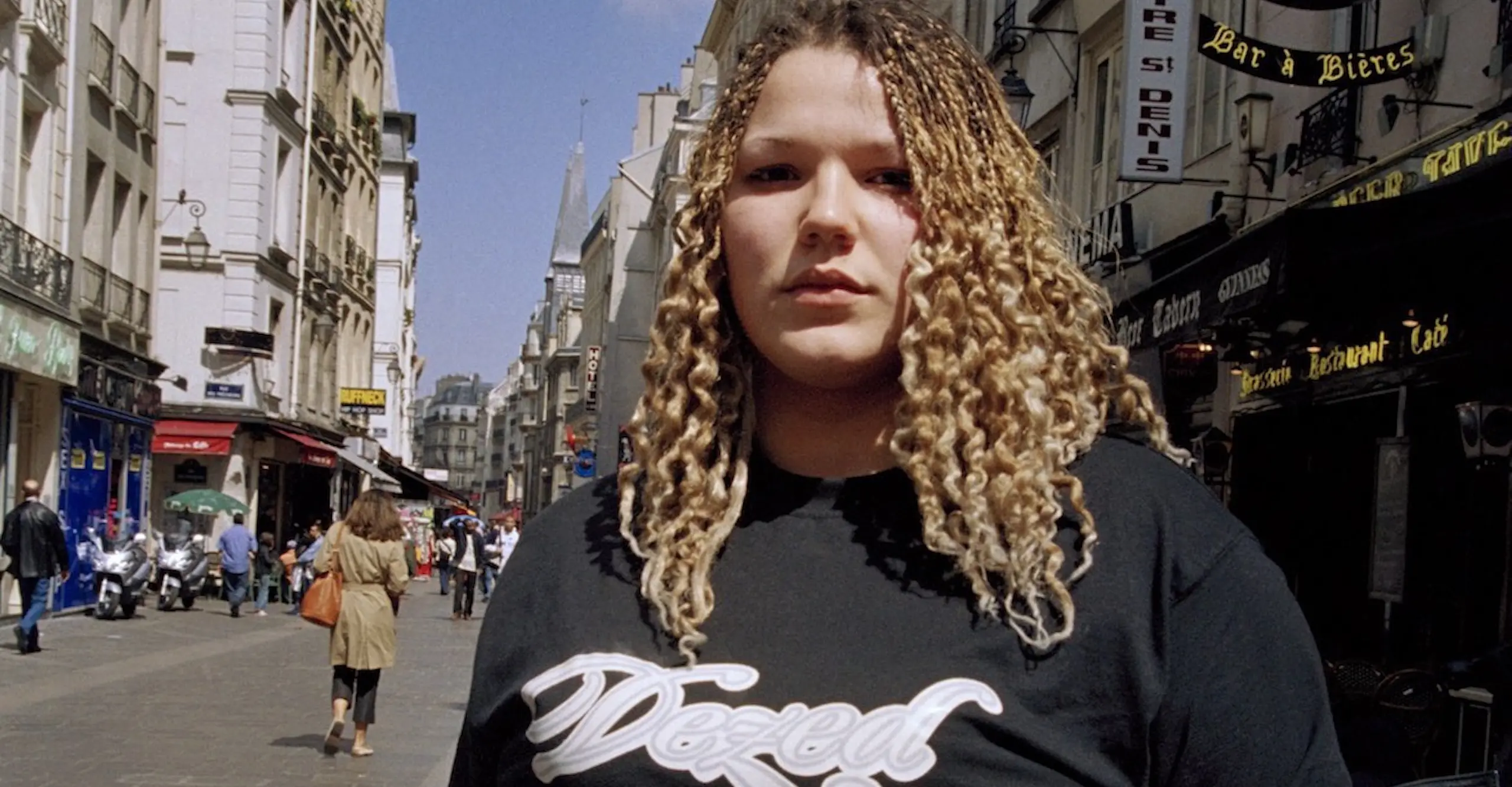 Head and shoulders of woman in black t-shirt and long curly blonde hair facing camera on a shopping street.