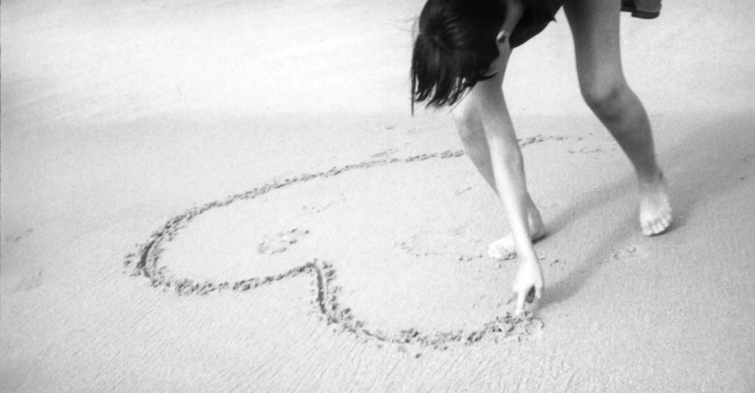 Black and white photograph of a the legs and arm of a child leaning over to drawa large heart in the sand.