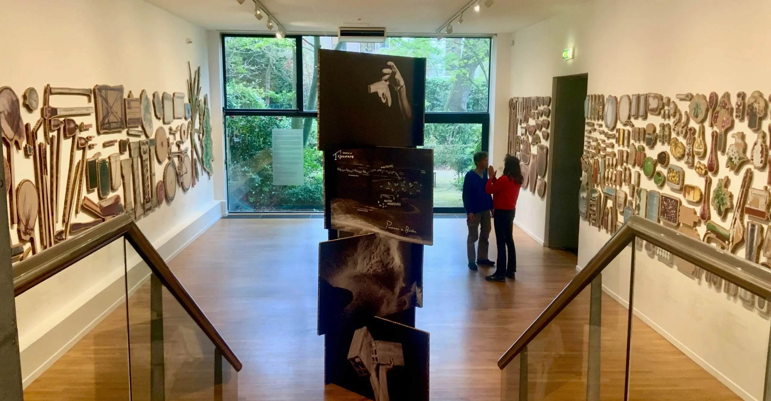 An exhibtion room with a large glass window on the far side. Photographs are stacked in a column in the middle of the room, objects printed on wood hang on the wall 