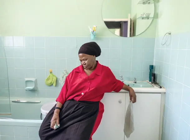 Colour photograph of a woman sat in a bathroom looking down. She holds a sponge in her hand.